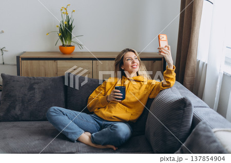 Happy woman taking selfie with smartphone on sofa holding coffee in cozy home interior with natural light and relaxed lifestyle vibe 137854904