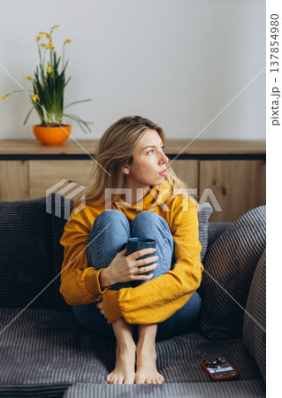 Lonely sad woman sitting on sofa holding coffee feeling anxiety and depression in quiet home interior with soft natural light Lonely sad woman sitting on sofa holding coffee feeling anxiety and depression in quiet home interior with soft natural light 137854980