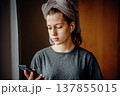 Teen girl standing by the window and looking at her smartphone, with earphones and a towel on her head. Natural home scene about communication, music, morning routine and everyday life. 137855015