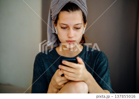 Portrait of a teen girl at home with a smartphone in her hands, earphones and a towel on her head. Natural scene about reading a message, screen focus, music and a quiet personal moment. Portrait of a teen girl at home with a smartphone in her hands, earphones and a towel on her head. Natural scene about reading a message, screen focus, music and a quiet personal moment. 137855016