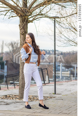 Elegant young woman in old money style walking along riverside promenade near pier in Prague during early spring. Understated luxury and timeless European lifestyle concept. Elegant young woman in old money style walking along riverside promenade near pier in Prague during early spring. Understated luxury and timeless European lifestyle concept. 137859085