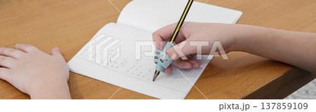 Child practicing handwriting with a special finger grip trainer. Close-up of kid's hand holding pencil correctly. Development of fine motor skills and calligraphy. Child practicing handwriting with a special finger grip trainer. Close-up of kid's hand holding pencil correctly. Development of fine motor skills and calligraphy. 137859109
