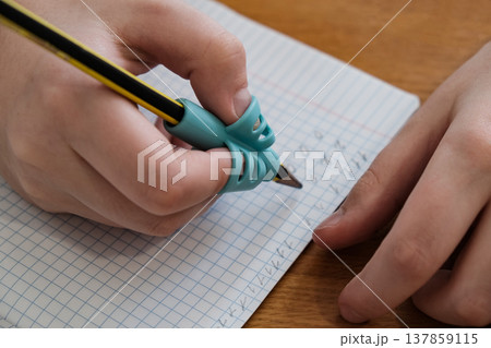 Child practicing handwriting with a special finger grip trainer. Close-up of kid's hand holding pencil correctly. Development of fine motor skills and calligraphy. 137859115
