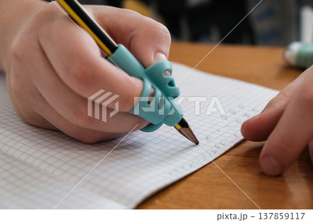 Child practicing handwriting with a special finger grip trainer. Close-up of kid's hand holding pencil correctly. Development of fine motor skills and calligraphy. 137859117