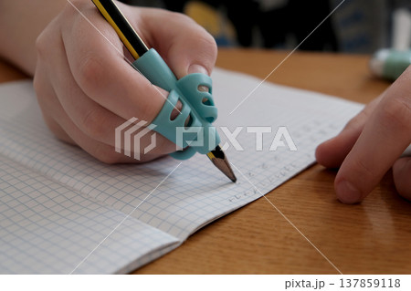 Child practicing handwriting with a special finger grip trainer. Close-up of kid's hand holding pencil correctly. Development of fine motor skills and calligraphy. 137859118