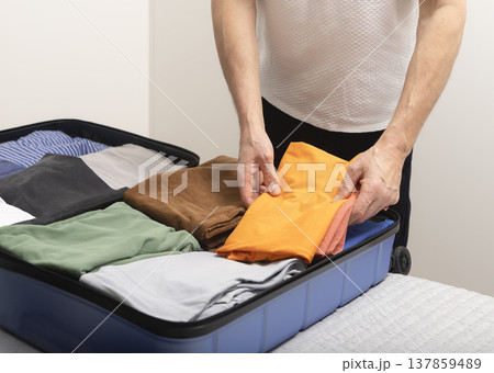 Weekend packing scene in an apartment with an open suitcase on the bed. Cropped male hands fold Weekend packing scene in an apartment with an open suitcase on the bed. Cropped male hands fold 137859489
