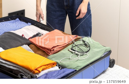Open suitcase lies on the bed filled with clothes and travel electronics. Cropped female hands place Open suitcase lies on the bed filled with clothes and travel electronics. Cropped female hands place 137859649