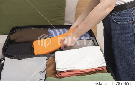 Open suitcase rests on the bed as clothes stack for an upcoming journey. Female hands fold essential Open suitcase rests on the bed as clothes stack for an upcoming journey. Female hands fold essential 137859659