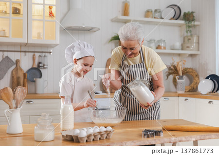 Happy family in kitchen. Grandmother and granddaughter child cook in kitchen together. Grandma teaching kid girl knead dough bake cookies. Household teamwork helping family generations concept 137861773