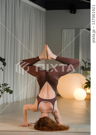 Woman practices handstand yoga pose in a studio with plants and soft lighting during a fitness session in the morning Woman practices handstand yoga pose in a studio with plants and soft lighting during a fitness session in the morning 137861921