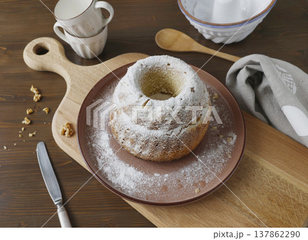 Gugelhupf bundt cake dusted with powdered sugar on plate. Rustic breakfast setting with wooden cutting board and kitchen utensils. Gugelhupf bundt cake dusted with powdered sugar on plate. Rustic breakfast setting with wooden cutting board and kitchen utensils. 137862290