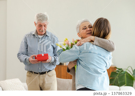 Happy family granddaughter gives gift box spring tulip flowers to grandparents. Teenage girl with bouquet surprise gift box getting congratulations on grandparents day. Family celebrating birthday Happy family granddaughter gives gift box spring tulip flowers to grandparents. Teenage girl with bouquet surprise gift box getting congratulations on grandparents day. Family celebrating birthday 137863329