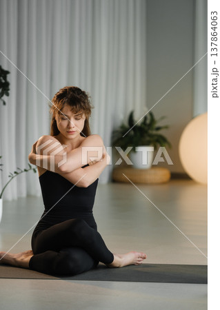 Woman practices yoga indoors while sitting on a mat in a well-lit studio with plants and soft lighting during a morning session Woman practices yoga indoors while sitting on a mat in a well-lit studio with plants and soft lighting during a morning session 137864063