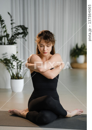 Woman practices yoga indoors while sitting on a mat in a well-lit studio with plants and soft lighting during a morning session Woman practices yoga indoors while sitting on a mat in a well-lit studio with plants and soft lighting during a morning session 137864065