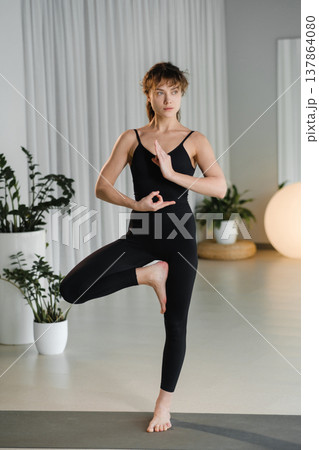 Woman practices yoga in a studio with plants and soft lighting during daytime, showing a balancing pose on one leg with arms raised 137864080