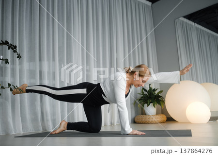 Woman practices yoga in a bright room with white curtains and indoor plants at an afternoon class focusing on balance and strength during a workout session Woman practices yoga in a bright room with white curtains and indoor plants at an afternoon class focusing on balance and strength during a workout session 137864276