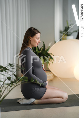 Pregnant woman in yoga pose performing meditation indoors during morning light in a calm space Pregnant woman in yoga pose performing meditation indoors during morning light in a calm space 137864369