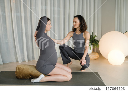 Pregnant woman stretches during a yoga session with instructor in a bright room filled with plants Pregnant woman stretches during a yoga session with instructor in a bright room filled with plants 137864398