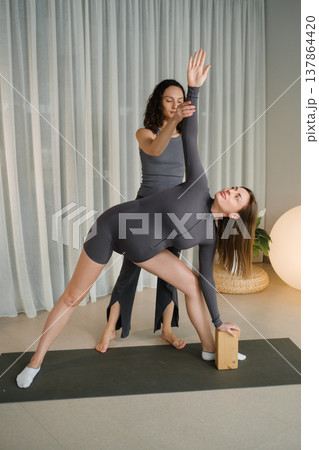 Yoga class takes place in a bright studio where two women practice a pose with support from a teacher and a yoga block 137864420