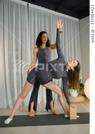 Yoga class takes place in a bright studio where two women practice a pose with support from a teacher and a yoga block 137864421