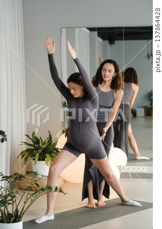Yoga practice takes place in a bright room with plants as an instructor assists a woman who is pregnant during a stretch at morning time Yoga practice takes place in a bright room with plants as an instructor assists a woman who is pregnant during a stretch at morning time 137864428