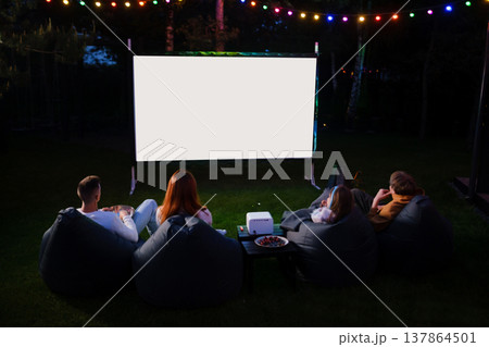 Friends enjoy movie night in backyard under string lights with a projector and bean bags at dusk 137864501