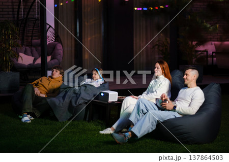 People enjoy movie night outdoors in their backyard using a projector while sitting on bean bag chairs under string lights during the evening People enjoy movie night outdoors in their backyard using a projector while sitting on bean bag chairs under string lights during the evening 137864503