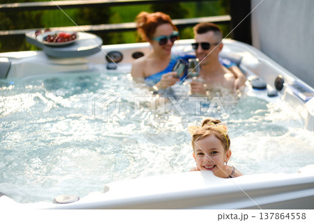 Family enjoys time together in hot tub while girl plays and parents relax on sunny day in backyard Family enjoys time together in hot tub while girl plays and parents relax on sunny day in backyard 137864558