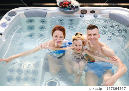 Family enjoys time together in hot tub during sunny day in backyard setting with snacks on table nearby Family enjoys time together in hot tub during sunny day in backyard setting with snacks on table nearby 137864567