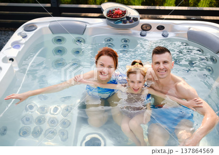 Family enjoys time together in hot tub during sunny day in backyard setting with snacks on table nearby Family enjoys time together in hot tub during sunny day in backyard setting with snacks on table nearby 137864569