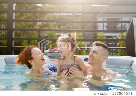 Family enjoys time together in hot tub during afternoon at home while laughing and playing with daughter in warm weather, creating happy moments and memories Family enjoys time together in hot tub during afternoon at home while laughing and playing with daughter in warm weather, creating happy moments and memories 137864571