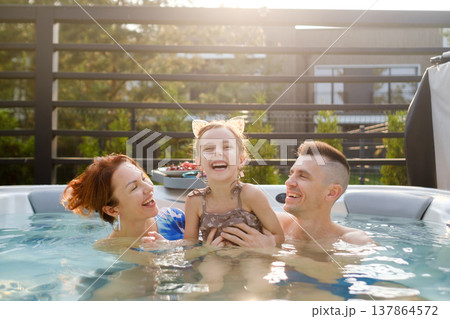 Family enjoys time in a hot tub with a child laughing in sunny weather in a backyard setting near trees and plants in the afternoon Family enjoys time in a hot tub with a child laughing in sunny weather in a backyard setting near trees and plants in the afternoon 137864572