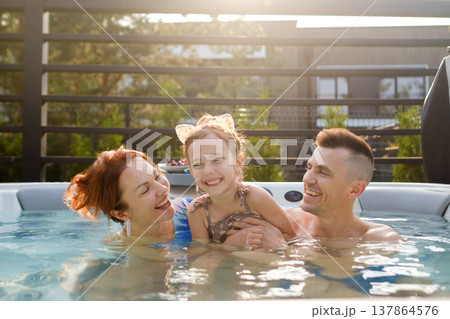 Family enjoys time together in a hot tub during a sunny afternoon in their backyard, sharing smiles and laughter with their daughter after a long day 137864576