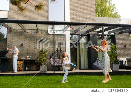 Family plays frisbee in backyard during sunny afternoon while a man cooks nearby Family plays frisbee in backyard during sunny afternoon while a man cooks nearby 137864580