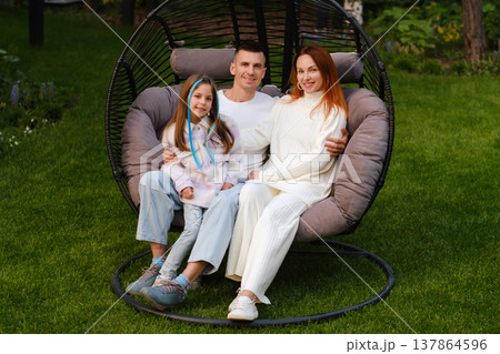 Family enjoys time together in a cozy outdoor chair at a green garden on a sunny day while sharing smiles and laughter Family enjoys time together in a cozy outdoor chair at a green garden on a sunny day while sharing smiles and laughter 137864596