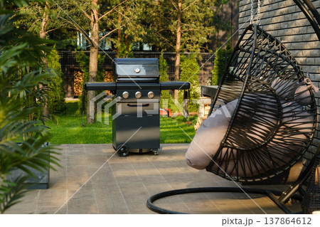 Grill stands on patio next to hanging chair in green backyard during sunny afternoon Grill stands on patio next to hanging chair in green backyard during sunny afternoon 137864612