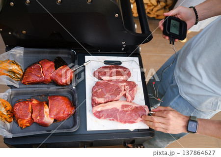 Grilling meat and checking temperature at outdoor barbecue event in backyard on sunny day with friends enjoying food preparation Grilling meat and checking temperature at outdoor barbecue event in backyard on sunny day with friends enjoying food preparation 137864627