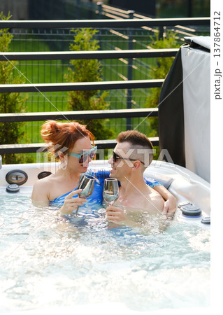 Couple enjoys sparkling drinks in hot tub during sunny day in backyard setting surrounded by greenery Couple enjoys sparkling drinks in hot tub during sunny day in backyard setting surrounded by greenery 137864712
