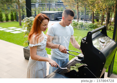 Couple cooking asparagus on grill during summer afternoon in backyard with green plants around and hot tub in background Couple cooking asparagus on grill during summer afternoon in backyard with green plants around and hot tub in background 137864728