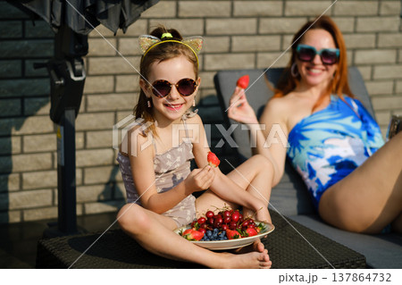 Happy moments at a summer gathering with a girl enjoying fresh fruits while a woman relaxes nearby in a comfortable setting 137864732