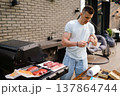 Man prepares meat for grilling at home backyard during afternoon with grill and table set up nearby 137864744