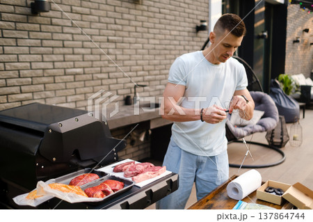 Man prepares meat for grilling at home backyard during afternoon with grill and table set up nearby 137864744