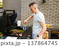 Cooking meat and vegetables on barbecue grill in backyard during sunny afternoon with friends nearby enjoying food and drinks 137864745
