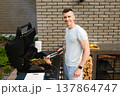 Man stands by a grill preparing meat and vegetables in a backyard setting during daytime with a brick wall as background 137864747