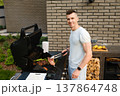 Man cooks meat on a grill in a backyard during a sunny day while smiling at the camera 137864748