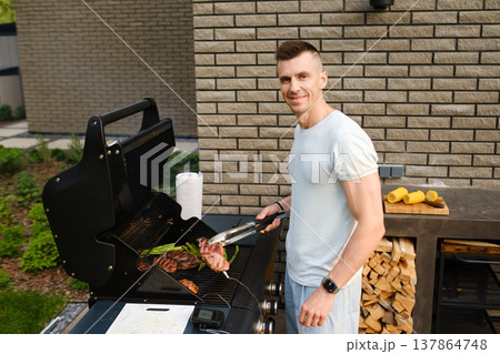 Man cooks meat on a grill in a backyard during a sunny day while smiling at the camera 137864748