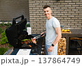 Man cooks meat on a grill in a backyard during a sunny day while smiling at the camera 137864749