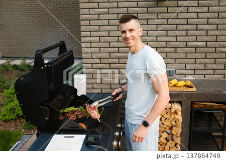 Man cooks meat on a grill in a backyard during a sunny day while smiling at the camera Man cooks meat on a grill in a backyard during a sunny day while smiling at the camera 137864749