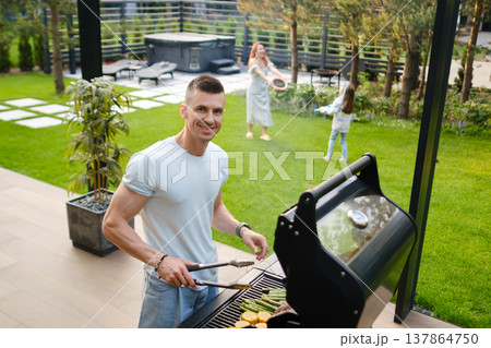 Man cooks on grill while friends enjoy time in yard during sunny day in backyard Man cooks on grill while friends enjoy time in yard during sunny day in backyard 137864750