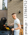 Man cooks meat and vegetables on a grill outside a house during daytime in a backyard 137864752